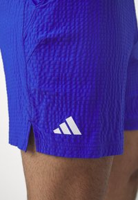 Blauwe, gestructureerde sportbroek met zijslitsen en het witte Adidas-logo. Het materiaal lijkt lichtgewicht en ademend, met een geometrisch patroon.