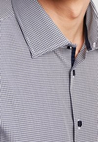 Kostkovaná košile s bílým a tmavě modrým vzorem, zapínání na knoflíky a kontrastním detailem vnitřního límce. Materiál se zdá být hladký a lehký.