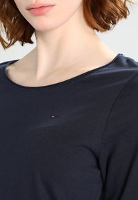 Námořnická modrá bavlněná košile s kulatým výstřihem, na levé straně hrudi s malým vyšitým logem v červené, bílé a modré barvě.