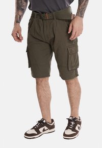 Pantaloni cargo in tessuto verde oliva, dotati di molteplici tasche laterali, una cintura con un accento marrone e un fitting rilassato. Indossati con sneakers marroni.