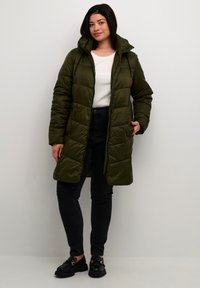 Donkergroene pufferjas met een capuchon, voorzien van horizontale stiksels, een rits aan de voorkant en twee zijzakken, gecombineerd met zwarte jeans en schoenen.