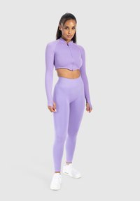 Haut zippé raccourci violet clair et legging taille haute assorti, en tissu lisse et élastique, associé à des chaussures de sport blanches.