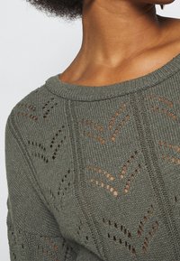 Pull en maille vert olive pâle avec un col rond, présentant un motif en maille ajourée et une texture côtelée sur les bords.