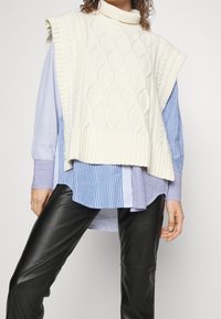 Béžová pletená tunika s kabelovým vzorem a vysokým výstřihem, vrstvená přes modrou pruhovanou košili a černé kožené kalhoty.