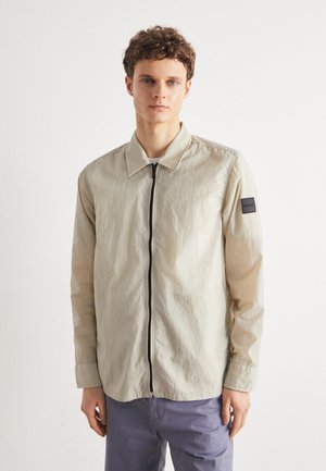Light beige lightweight jacket with a front zipper, collar, long sleeves, and a black logo patch on the left arm. Smooth texture.