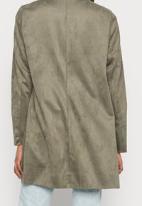 Vista posteriore di una persona che indossa una giacca in suede verde oliva larga e jeans azzurri su uno sfondo semplice.
