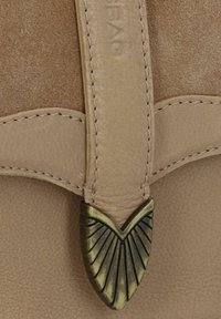 Dettaglio di una borsa in pelle beige che mostra una superficie marrone texturizzata, una tracolla liscia e un accento metallico a forma di V con scanalature.