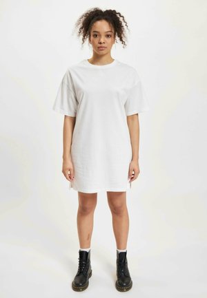 Witte katoenen T-shirt jurk met korte mouwen, voorzien van zijsplitten. Combinatie met zwarte veterschoenen en witte sokken. Zachte textuur.