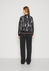 Veste bomber noire avec un motif tie-dye blanc, des finitions contrastantes, des poignets et un ourlet côtelés. Portée avec un pantalon en denim foncé taille haute.
