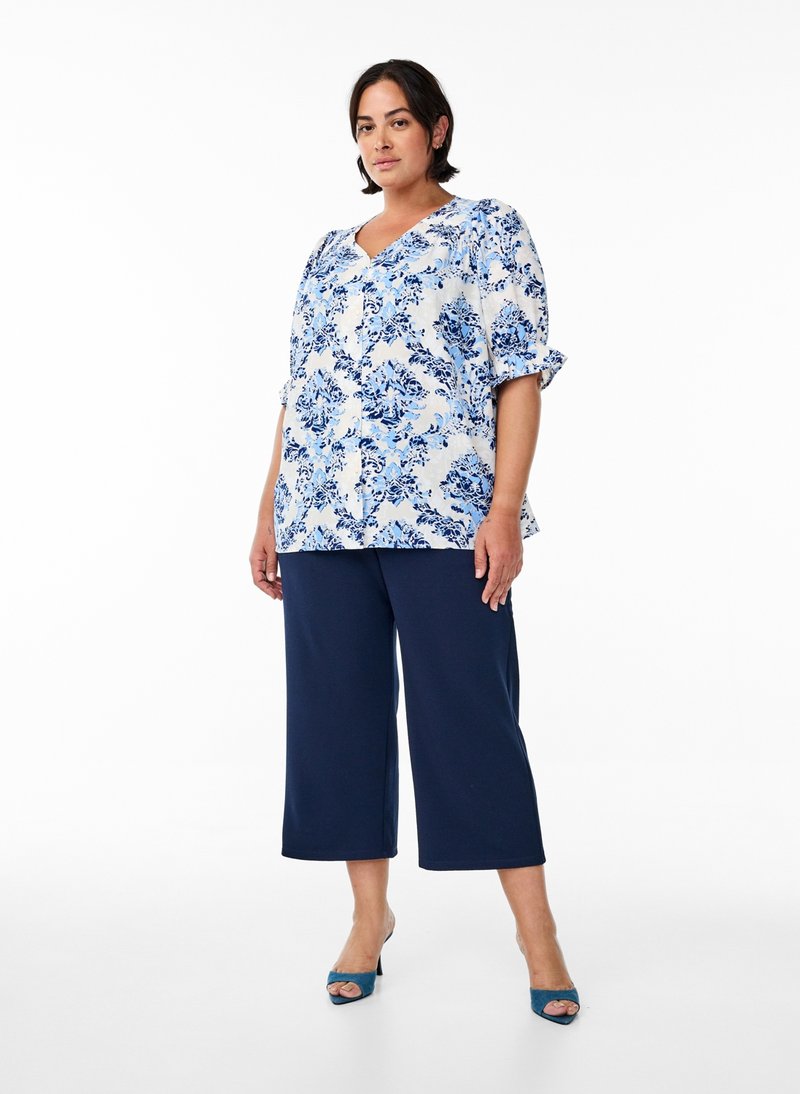 Mujer con blusa floral azul y blanca, pantalones anchos hasta la pantorrilla de color azul marino y tacones azules abiertos en los dedos, de pie frente a un fondo blanco.