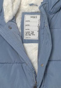 Ljusblå puffjacka med mjukt vitt fleecefoder, dragkedja fram, metallknappar och en inre etikett för personalisering.