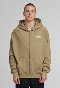 Beige zip-up hoodie with a kangaroo pocket, ribbed cuffs, and a logo on the left chest. Soft fabric with a smooth texture.