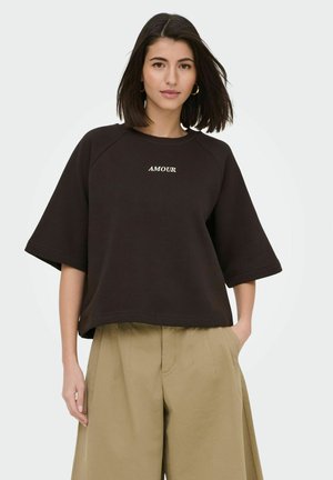 Kobieta o ciemnych włosach do ramion nosi czarny, oversize'owy top z krótkim rękawem z napisem "AMOUR" i luźne beżowe spodnie, pozując z jedną ręką w kieszeni.