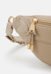Coin de sac en cuir beige texturé avec fermeture éclair dorée, tirettes en cuir tressé et détail en cuir cousu en forme de cœur près de la fixation de la bandoulière.