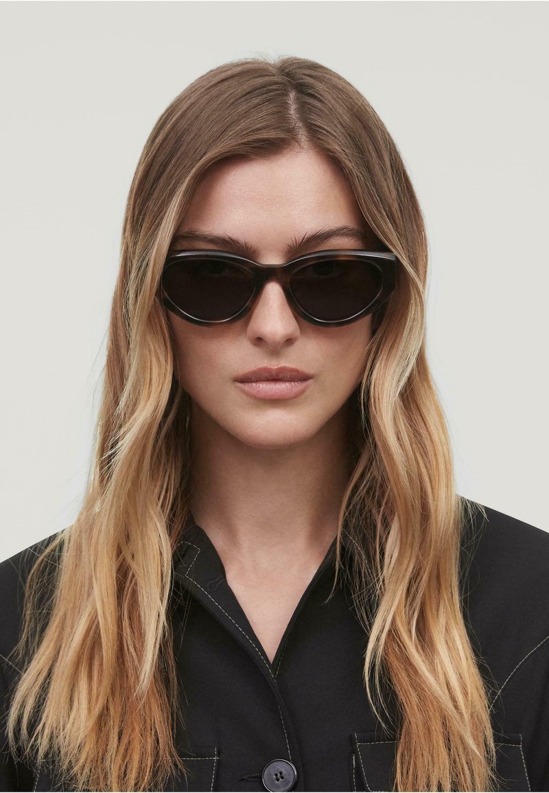 Gafas de sol de cat-eye negras con lentes oscuros, que presentan un acabado brillante y un marco elegante y angular. Llevadas por una modelo con el cabello largo y liso.