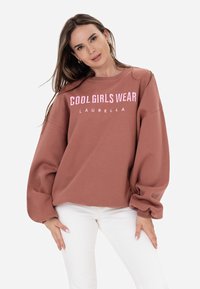 Rūsas krāsas džemperis ar puff piedurknēm, ar uzrakstu "COOL GIRLS WEAR LAURELLA" rozā krāsā. Valkāts kopā ar baltām biksēm.