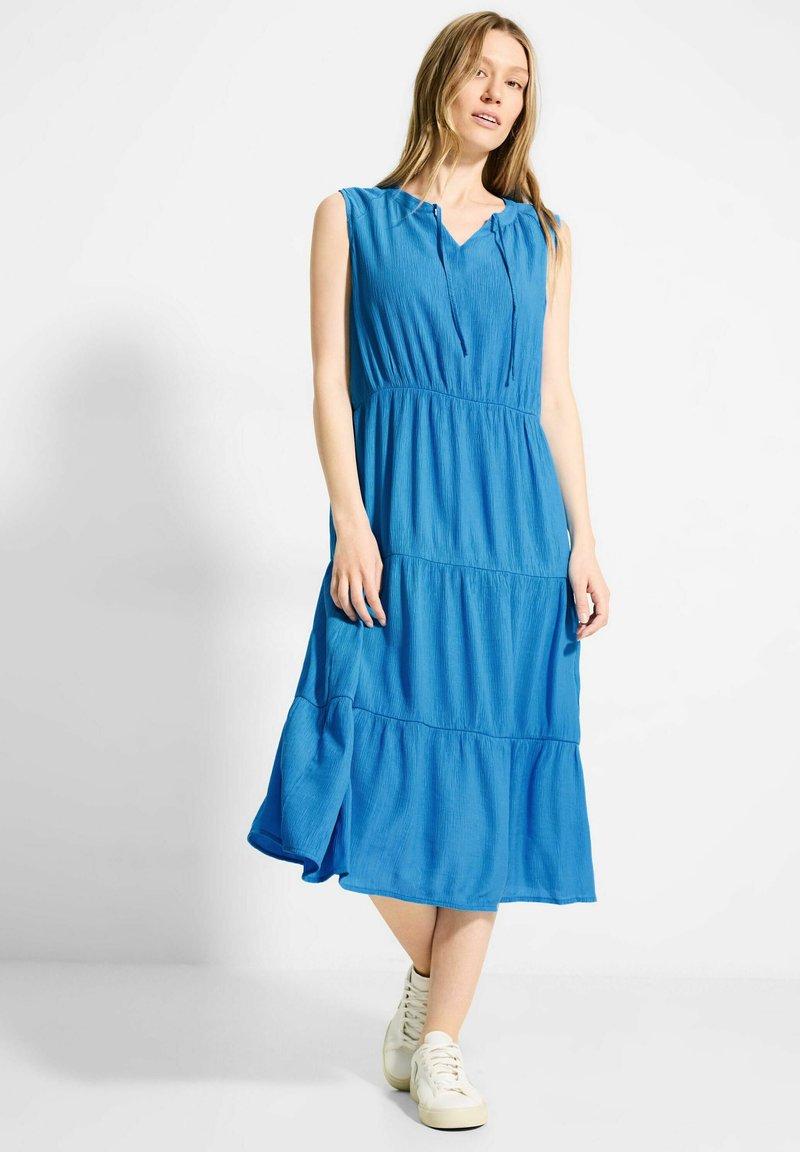 Blaues ärmelloses Kleid mit V-Ausschnitt, gestuftem Design und Kordelzugdetails. Hergestellt aus leichtem Stoff, kombiniert mit weißen Sneakers.