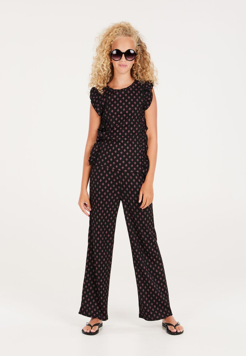 Zwarte jumpsuit met ruches aan de mouwen en een roze patroon. Wijde pijpen, gecombineerd met zwarte slippers en grote ronde zonnebril.