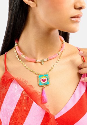 Colliers superposés comprenant un collier ras du cou en perles roses, une chaîne dorée ornée d'une fleur, et un pendentif carré coloré avec un pompon rose.