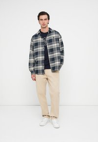 Hombre joven de pie con las manos relajadas, vistiendo una camisa de cuadros azul marino sobre una camiseta gráfica oscura, pantalones beiges y zapatillas blancas, contra un fondo blanco.