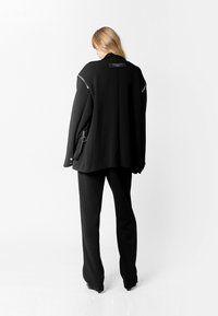 Chaqueta negra de gran tamaño con collar alto, detalles de cremallera plateada y bolsillos laterales, combinada con pantalones negros ajustados. Textura suave.