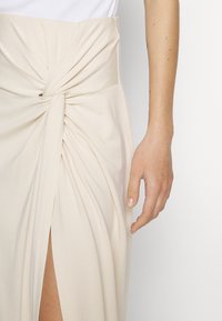 Jupe beige avec un détail noeud à l'avant et une fente haute sur la cuisse, portée avec un haut blanc et un bras nu visible côté droit.