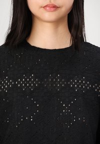 Top nero in tessuto testurizzato con intricate motivazioni a diamante e punti, dotato di scollatura rotonda. Pelle visibile attraverso il design intrecciato del tessuto.