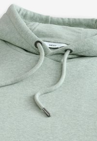 Lichtgroene hoodie met een gestructureerde stof. Voorzien van een capuchon met trekkoord en metalen uiteinden, en een label met de tekst "NEXT" aan de binnenkant van de halslijn.