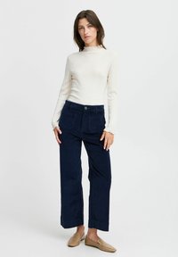 Pantalones de pana de pierna ancha en color azul marino, con bolsillos frontales y cierre de botón, combinados con una blusa de manga larga en color crema con textura de canalé y un cuello con volantes.