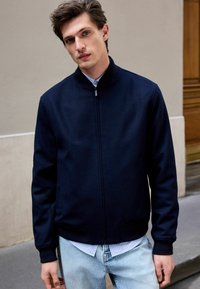 Jūrų laivyno zip-up striukė su aukštu apykakle, pagaminta iš lygaus audinio. Dėvima virš šviesiai mėlynos dryžuotos marškinėlės ir derinama su išblukusiomis džinsomis.