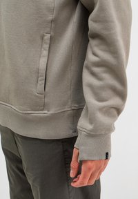 Hellgrauer Sweatshirt mit gerippten Bündchen und Saum, ausgestattet mit einer Seitentasche und weicher Textur. Über einer dunklen Hose getragen, mit einem Etikett am Bündchen.