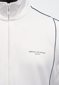 Biała kurtka zapinana na zamek, wykonana z gładkiego materiału, z wysokim kołnierzem, czarnymi lamówkami po bokach oraz małym logo na klatce piersiowej z napisem "ARMANI EXCHANGE MILANO."