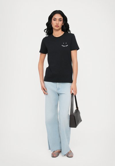 Black cotton t-shirt with a round neck and small white text. Paired with light blue flared jeans and multicolored striped sandals. Black handbag.