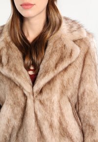 Hladký kožich z umělé kožešiny ve světlé hnědé barvě. Má široký límec a volnější střih. Nosený přes vínové tričko.