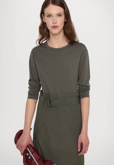 Abito verde oliva con maniche lunghe, scollatura rotonda e vita con cintura. Il modello tiene una borsa bordeaux. Tessuto liscio con un design semplice.
