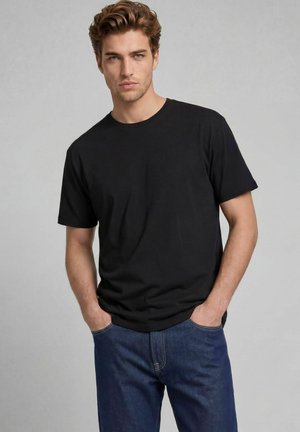 Jeune homme aux cheveux châtain clair portant un t-shirt noir uni à manches courtes et un jean bleu, les mains dans les poches, fond gris neutre.