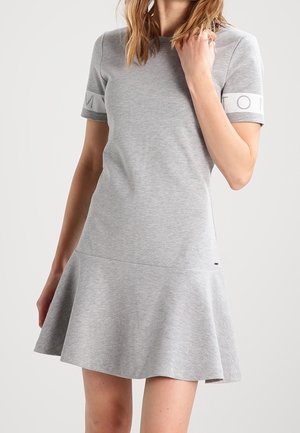 Mujer con vestido gris claro de manga corta, falda acampanada y detalle de texto en las mangas sobre un fondo liso.