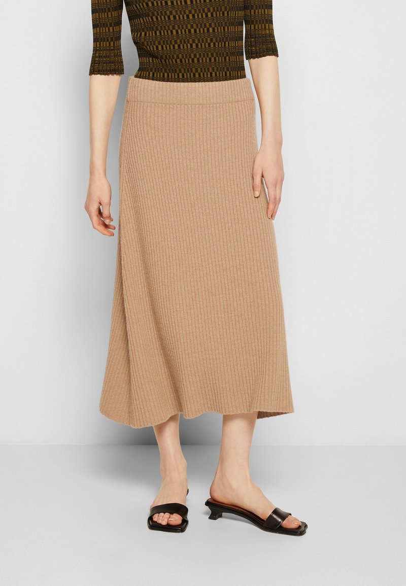 Femme portant une jupe midi beige côtelée, des sandales noires à petit talon et un haut marron à motifs à manches trois-quarts sur un fond uni.