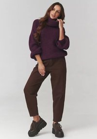 Fioletowy, oversize'owy sweter z wysokim kołnierzem, w połączeniu z brązowymi, zwężającymi się spodniami i masywnymi, ciemnymi butami, tworzy luźną sylwetkę.