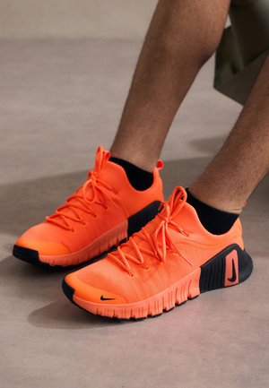 Stopy w jaskrawo pomarańczowych i czarnych sportowych trampkach z sznurowadłami, na gładkiej szarej powierzchni.