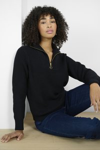 Pull en tricot noir avec un design à demi fermeture éclair et un accent de fermeture éclair doré. Assorti à un jean bleu foncé, assis contre un fond neutre.