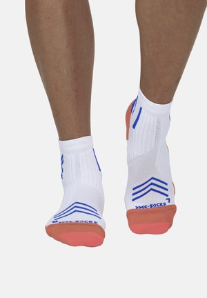 Piernas con calcetines tobilleros blancos con rayas azules y puntas naranjas, de pie sobre un fondo gris claro liso.