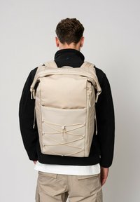 Sac à dos beige avec un tissu texturé, fermeture éclair, bretelles réglables et détail de cordon élastique à l'avant. Vue arrière sur fond neutre.