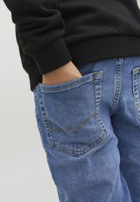 Jeans de mezclilla en un color azul claro, con costuras destacadas y dobles bolsillos traseros. También se muestra un puño de sudadera negra.