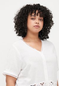 White, short-sleeve blouse with a V-neck and decorative lace trim. Textured fabric, featuring button front and lace detailing at the bottom.