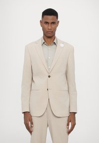 Un completo in lino beige presenta un blazer strutturato con due tasche frontali, bottoni marroni e una spilla da giacca a forma di fiore bianco, abbinato a una camicia verde chiaro.