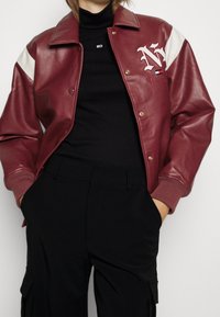 Burgunderrote Lederjacke mit weißen Schulterakzenten, Druckknöpfen und gesticktem Logo. Kombiniert mit einem schwarzen Rollkragenpullover und schwarzen Hosen.