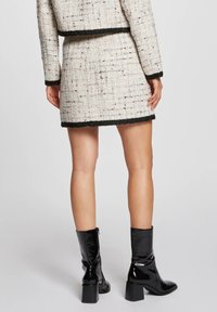 Conjunto de chaqueta y falda de tweed en crema con acentos en negro. La ropa presenta un patrón texturizado y se combina con botas de charol negras hasta la rodilla.