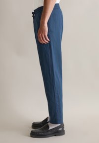 Marineblauwe broek in een lichte stof, met een elastische tailleband en een trekkoord. Gecombineerd met zwarte instappers en grijze sokken.