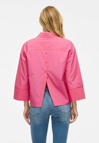 Camicia rosa a maniche corte con colletto, dettaglio di bottoni sul retro e design a spacco. Realizzata in un tessuto morbido e leggero.